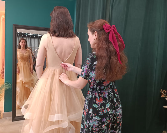 Daniella, la gérante du showroom accompagnant une cliente lors de son essayage de robes de soirée, en lui fermant la tirette dans le dos. La cliente s'admire dans le grand miroir face à elle. C'est l'expérience shopping du showroom privatisé, à Dijon.